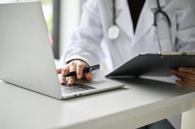 Cropped image of a professional female doctor in uniform working at her desk, using laptop and examining medical cases on clipboard.