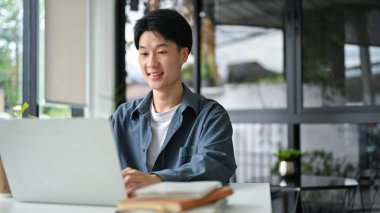Smart young Asian man in casual clothes looking at laptop screen, using portable laptop, remote working at the cafe.