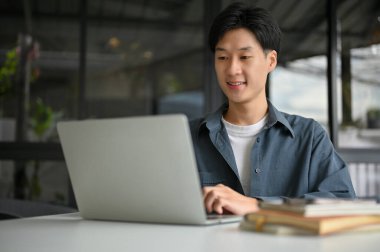 Smart and handsome young Asian male office worker or college student remote working at the coffee shop, using laptop and focusing on his project.