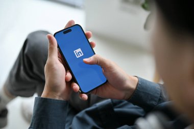 Chiang Mai, Thailand - Feb 17 2023: close-up image of an Asian man using LinkedIn on his smartphone. iPhone with LinkedIn logo on screen. Job and internship platform service