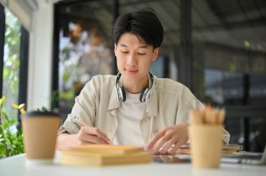Smart and focused young Asian male college student is in the coffee shop doing homework, reading a book, preparing for an exam.