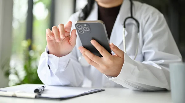 Cropped front view of a professional Asian female doctor in white gown using her smartphone to check her surgery\'s schedule or reading medical online articles at her desk.