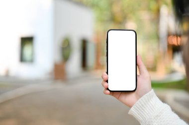 Close-up image of a woman's hand holding a smartphone white screen mockup over blurred background of beautiful cafe building.
