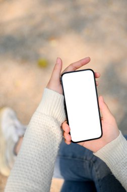 Close-up image of a woman's hand in white sweater holding a smartphone white screen mockup over blurred street in background.