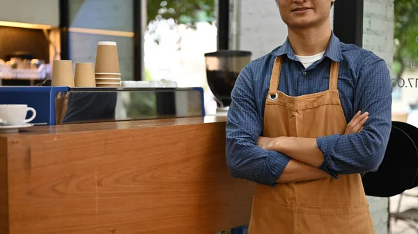 Successful Asian male barista or waiter in apron stands with arms crossed in front of the cashier counter in the coffee shop or cafe. cropped image. SME small business concept