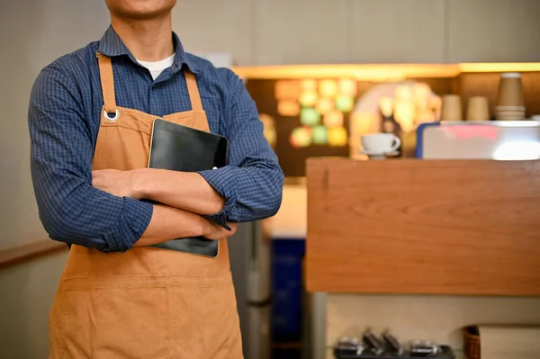 Confident Asian male barista or waiter in apron stands with arms crossed in the coffee shop or restaurant. cropped image