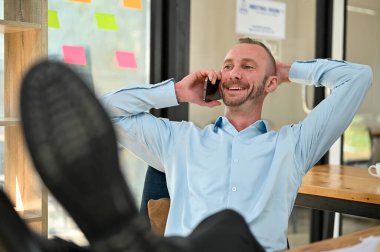 Handsome and happy Caucasian male engineer putting his legs on table and hand behind head while talking on the phone with client and chilling in the office.