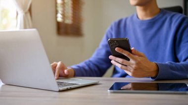Cropped image of a smart Asian male college student on cozy sweater using laptop and smartphone at his desk. technology and people concept 