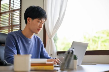 Smart young Asian male office worker or college student focusing on his project on laptop, working in his home office.