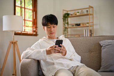 Handsome young Asian man in casual clothes using his smartphone, chatting with his friends on scrolling on social media while relaxing on sofa in his living room.