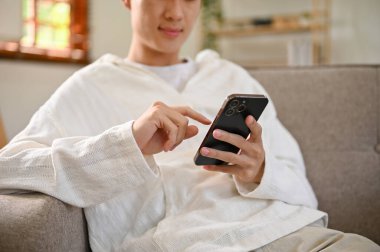 Cropped image of a young Asian man in casual clothes using his smartphone, chatting with his friends on scrolling on social media while relaxing on sofa in his living room.