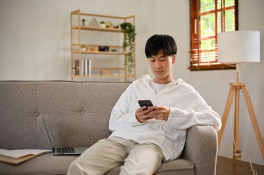 Handsome and relaxed young Asian man in casual clothes using his smartphone, chatting with his friends on scrolling on social media while relaxing on sofa in his living room.