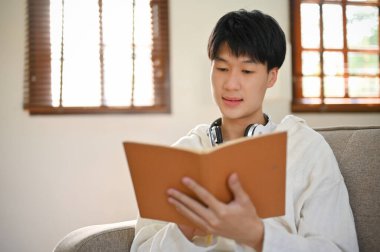 Smart and focused young Asian man reading a book on sofa in the living room. education and leisure concept