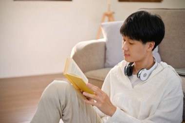 Happy and smart young Asian man reading a book in his minimal living room, enjoys reading novel or fiction. education concept