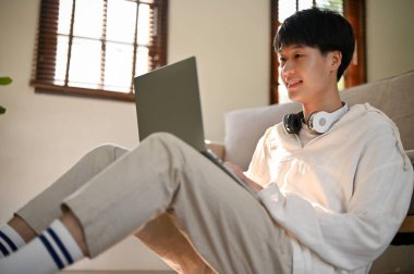 Happy and handsome young Asian man using laptop computer while relaxing in his living room. people an technology concept