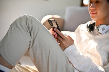 Cropped image of a handsome young Asian man with headphones using his smartphone while relaxing in his living room.