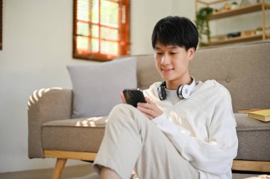 Positive young Asian man in comfy clothes is in the living room using his smartphone to chat with his friends.
