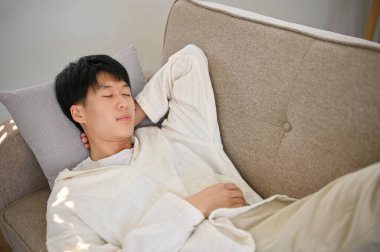 Top view of a relaxed and calm young Asian man in comfy clothes sleeping on the sofa in his living room. napping, laying, fall asleep, resting, snooze.