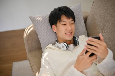 Top view of a happy handsome young Asian man in comfy clothes laying on sofa and using his smartphone.