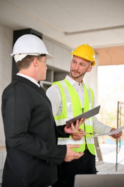 Portrait, Professional Caucasian male construction engineer manager in a yellow hardhat discussing with a businessman while inspecting the building together.