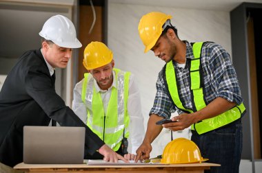 Professional Asian male foreman in uniform and yellow safety hat is talking and planning building process with a Caucasian businessman and construction inspector at the construction site.