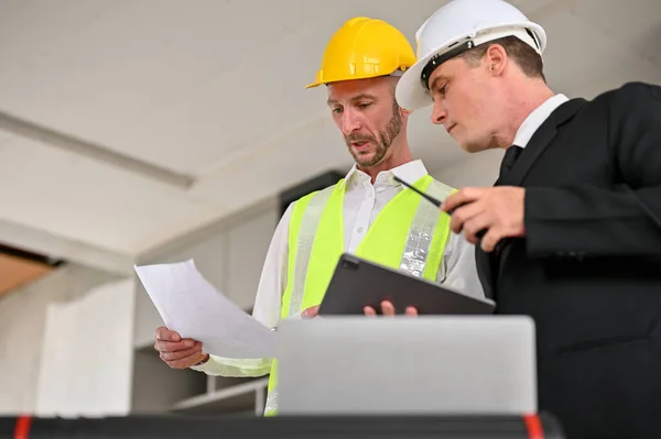 A professional Caucasian foreman explains the building process to a businessman while checking the building scale on a blueprint. Business and Industrial concept