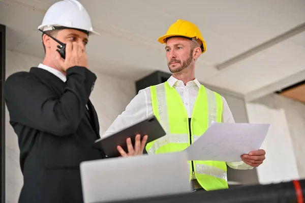Serious Caucasian male foreman or construction engineer manager meeting with a businessman at the construction site. A businessman using a walkie-talkie while meeting with a foreman.