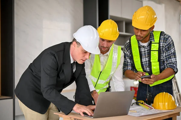 Professional Caucasian businessman in white hardhat and business suit using laptop while meeting and discussing the building plan with a team of construction engineers. Diverse workers concept