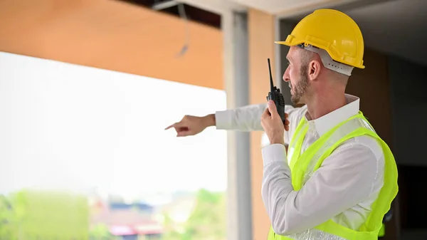 Serious and determined Caucasian male foreman or construction manager in a yellow hardhat using a walkie-talkie to command his construction workers. back view