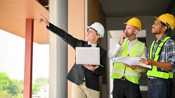 Successful Caucasian businessman in a white hardhat and business suit inspects the building at the site with a construction team. Diverse workers concept