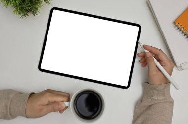 A female's hand holding a coffee cup and a stylus pen, working at her office desk, tablet white screen mockup on white tabletop. top view