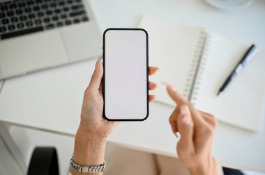 Top view of a modern smartphone white screen mockup is in a woman's hand over modern office desk.