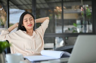 Beautiful and relaxed millennial Asian businesswoman or female manager stretching hands, eyes closed, taking a break from work at her desk.