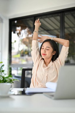 Beautiful millennial Asian businesswoman or female manager stretching hands, relaxing at her office desk.