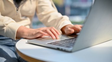 Cropped and close-up image of a young Asian woman in casual clothes using her laptop computer, typing on keyboard, working on her project.