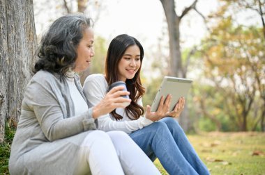 Mutlu ve nazik Asyalı büyükanne torunuyla yeşil parkta takılıyor, ağacın altında oturup tabletten komik videolar izliyor..