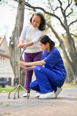 Asyalı kadın bakıcı ya da hemşire yaşlı bir bayana, yeşil parkta birlikte yürümesi için yardım ve destek ediyor..