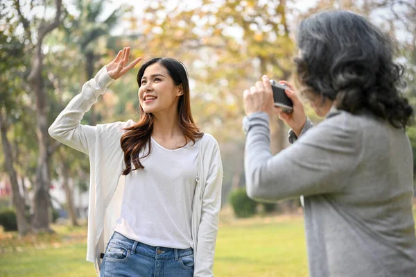 Happy Asian grandma taking a picture of her beautiful granddaughter with a retro camera while strolling in the green park together. Happy family and leisure concept
