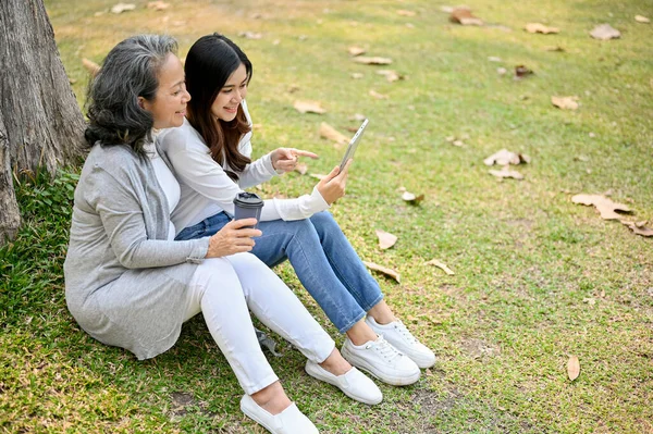 Lovely Asian granddaughter and grandmother sitting under the tree in the green park together, watching video on tablet, sipping coffee and chilling.