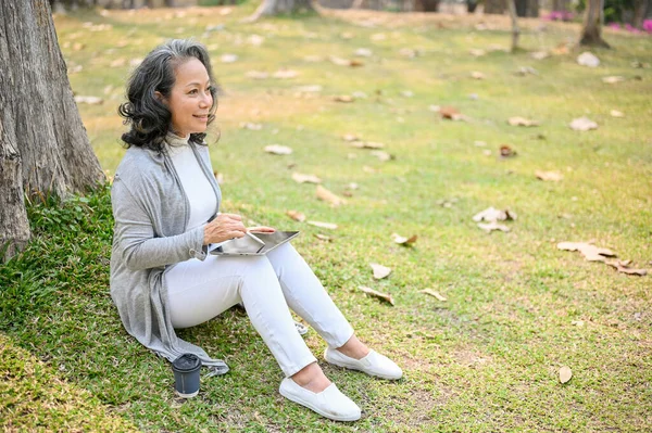 公園の木の下で草の上でリラックスして座っている間 彼女のデジタルタブレットを使用してカジュアルな服の幸せと魅力的な60歳のアジアの女性の側のビュー ライフスタイルのコンセプト — ストック写真