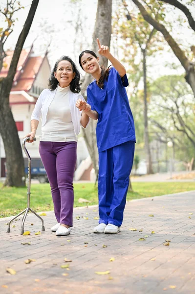 stock image Portrait of a caring Asian female caregiver or nurse assistant enjoys talking and walking in the green park with an old lady patient.