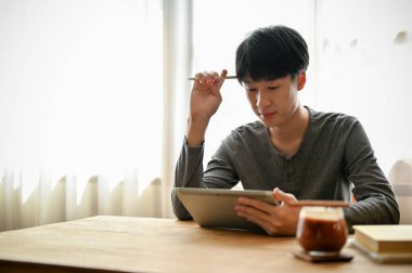 Yakışıklı ve düşünceli bir Asyalı. Günlük kıyafetler içinde. Okul projesini düşünüyor ve planlıyor. Dijital tablet kullanıyor, kafede oturuyor..