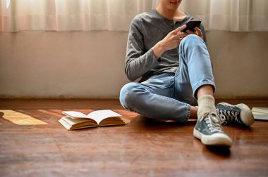 Moda kıyafetleri içinde yakışıklı bir Asyalı 'nın kırpılmış görüntüsü odasında yerde oturmuş telefonunda kayıyor. Yaşam tarzı ve teknoloji kavramı