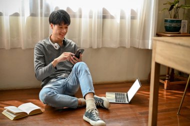 Moda kıyafetleri içinde yakışıklı ve mutlu Asyalı genç adam odasında yerde oturmuş telefonuyla oynaşıyor. Yaşam tarzı ve teknoloji kavramı