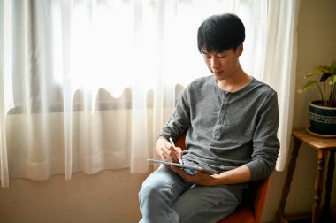 Güzel görünümlü Asyalı genç adam oturma odasının penceresinin yanındaki sandalyede otururken dijital tabletini kullanıyor..