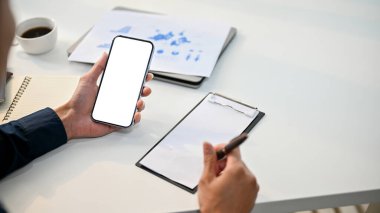 Profesyonel Asyalı iş adamının yakın plan görüntüsü telefonunu kullanıyor ve masasındaki kağıda bir şeyler yazıyor. akıllı telefon beyaz ekran modeli.