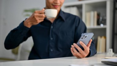 Asyalı başarılı bir iş adamının, masasında sabah kahvesi içerken telefonunu internet haberlerini okumak için kullanmasının kırpılmış ve seçici odak noktası görüntüsü..