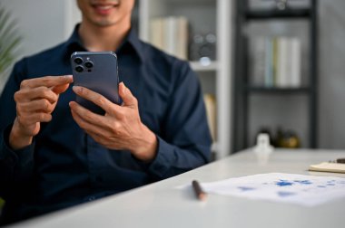 Bin yıllık mutlu Asyalı iş adamının ofisteki masasında telefonunu kullanırken çekilmiş görüntüsü..