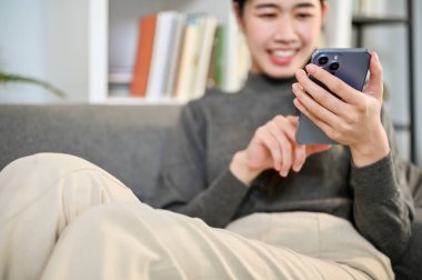 Kırpılmış görüntü, alımlı ve sıradan kıyafetleri içinde güzel bir Asyalı kadın telefonunu kullanıyor, oturma odasındaki kanepede dinlenirken sosyal medyada kayıyor..