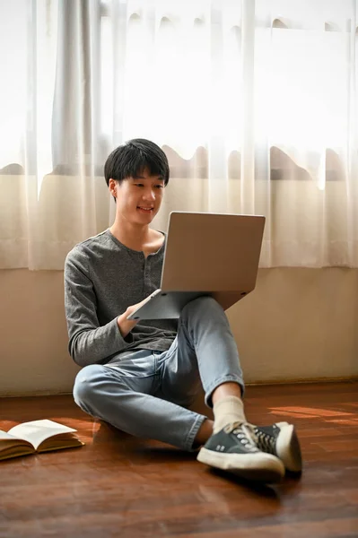 一个英俊而快乐的亚洲年轻人的画像坐在房间的地板上 用他的笔记本电脑来管理他的学业 — 图库照片
